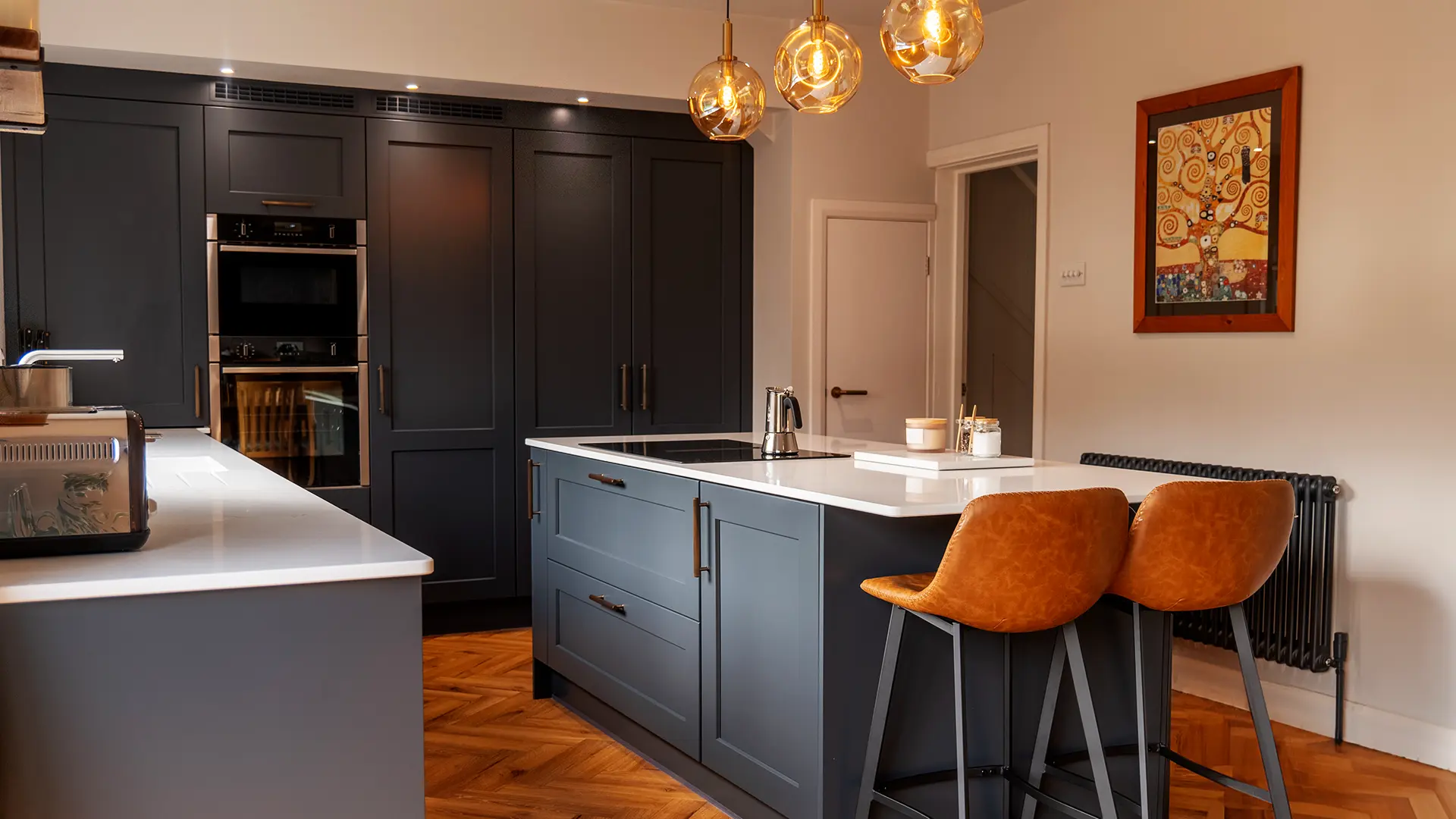Sophisticated dark navy shaker kitchen with leather bar stools, globe pendant lights, and parquet wooden flooring.