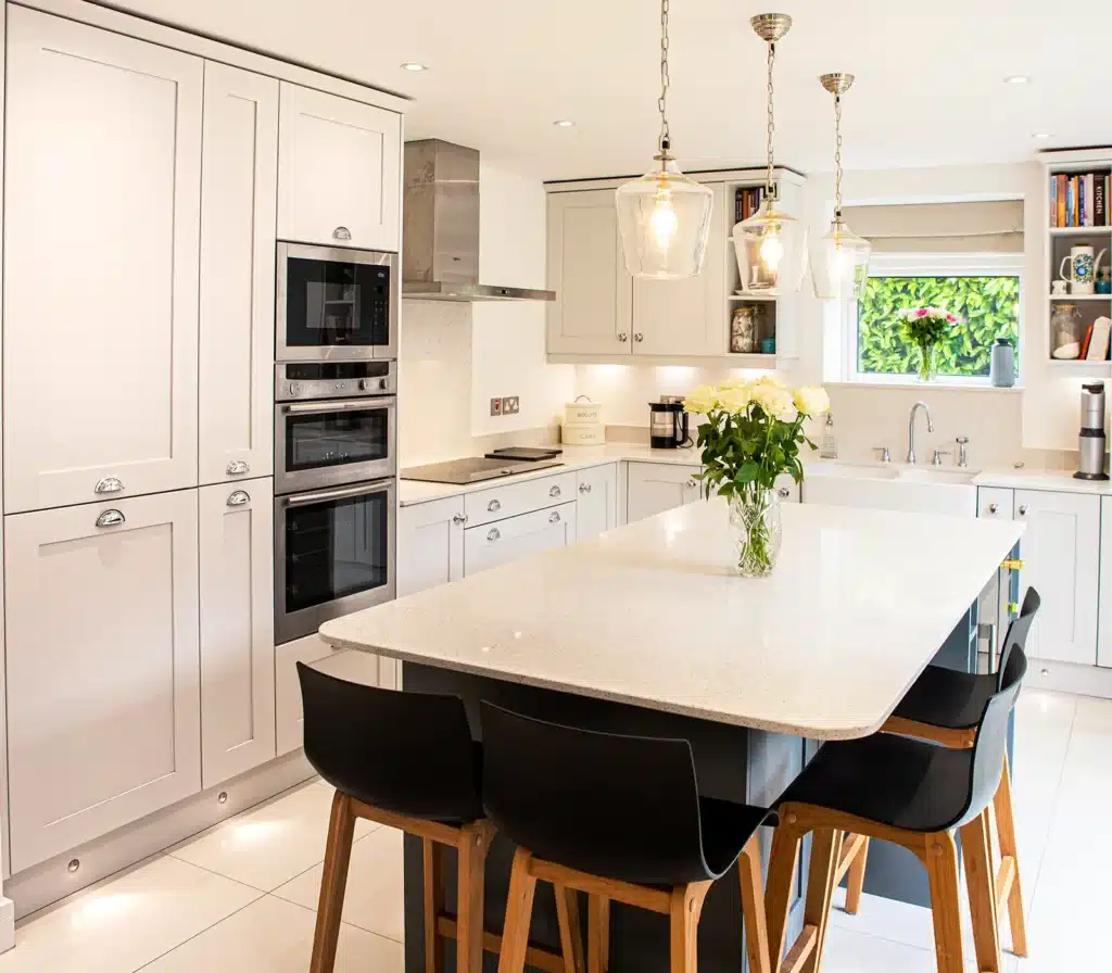 A bright Shaker kitchen with soft grey cabinets, a large quartz island, pendant lighting, integrated appliances, and a classic Belfast sink, styled with fresh flowers for a clean, modern look.