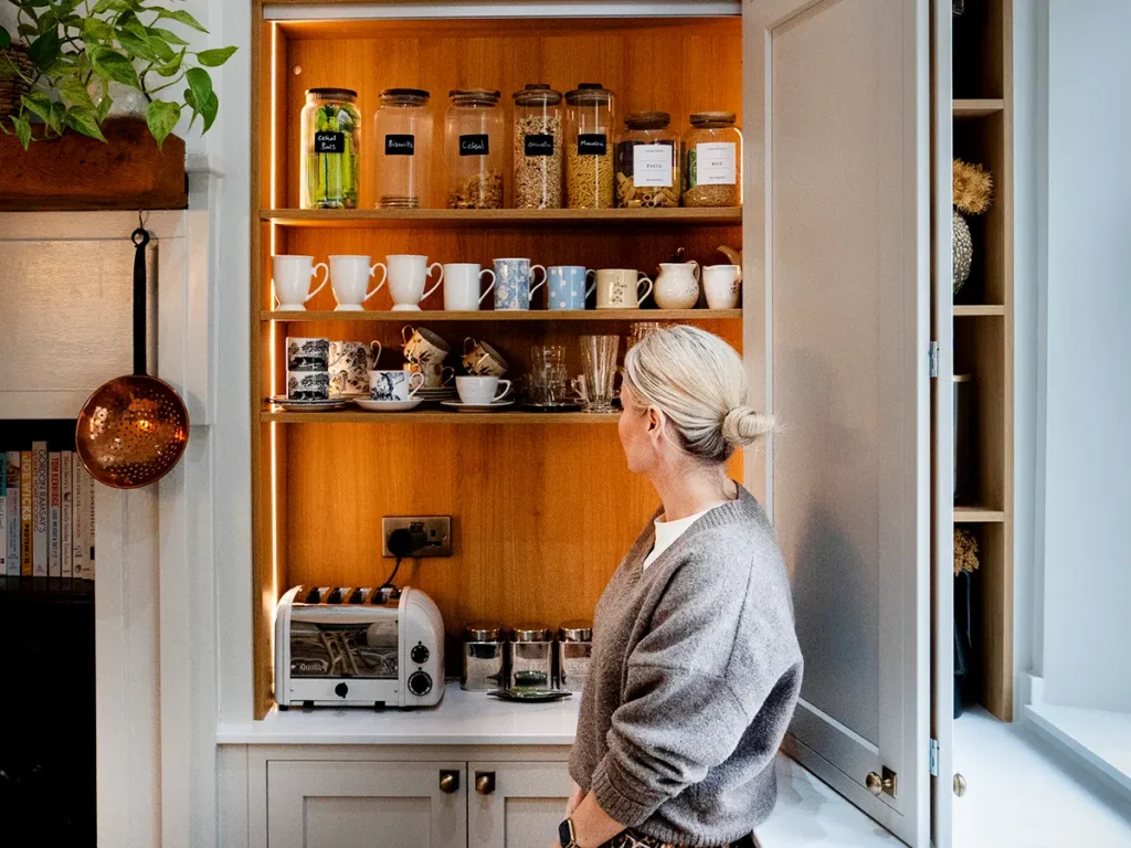 Bespoke breakfast pantry cabinet with warm wood interior shelving, integrated LED lighting, and organized glass storage jars, being used by a woman.