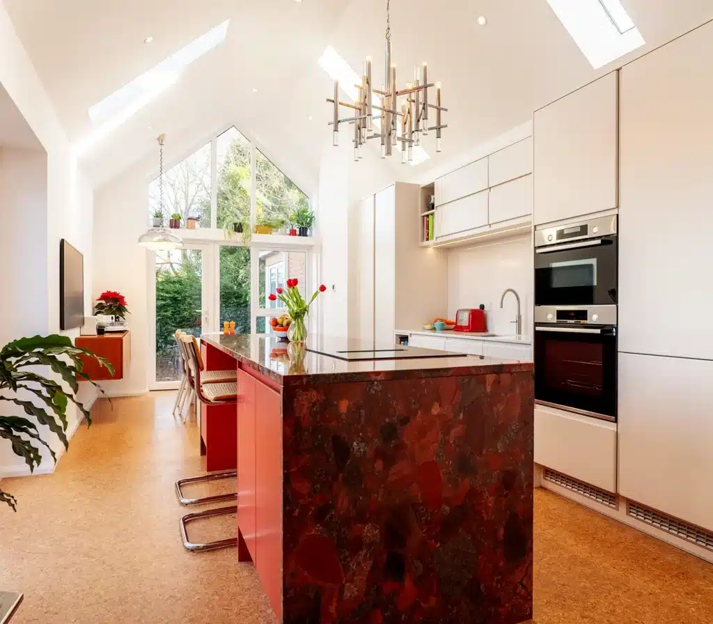 Modern bespoke kitchen with white handleless cabinets, red island, statement stone worktop, vaulted ceiling and large garden-facing windows.